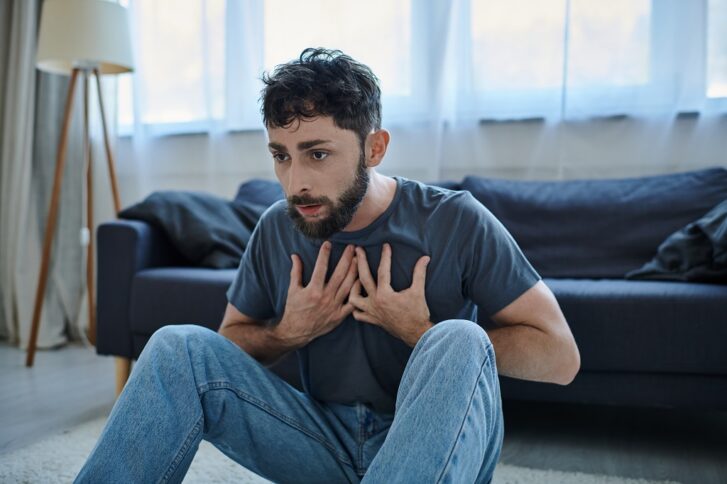 Man sitting on the floor holding his chest and struggling to breathe during a panic attack at home