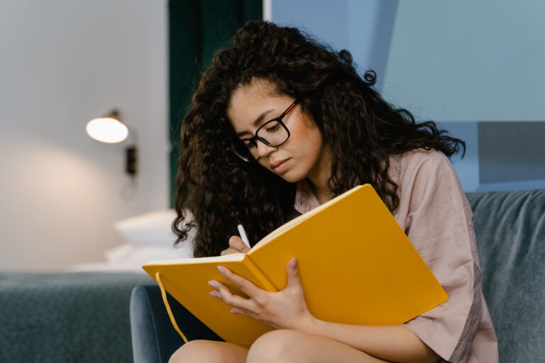 Young woman writing in a notebook at home as part of a reflective mental health self-care routine.